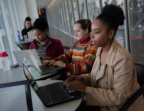 Three females sitting at small tables with computers in a hallway.